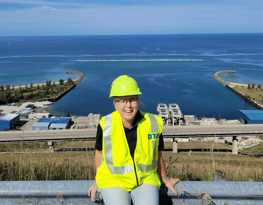 Woman in hard hat and safety gear with power plant and water in the background