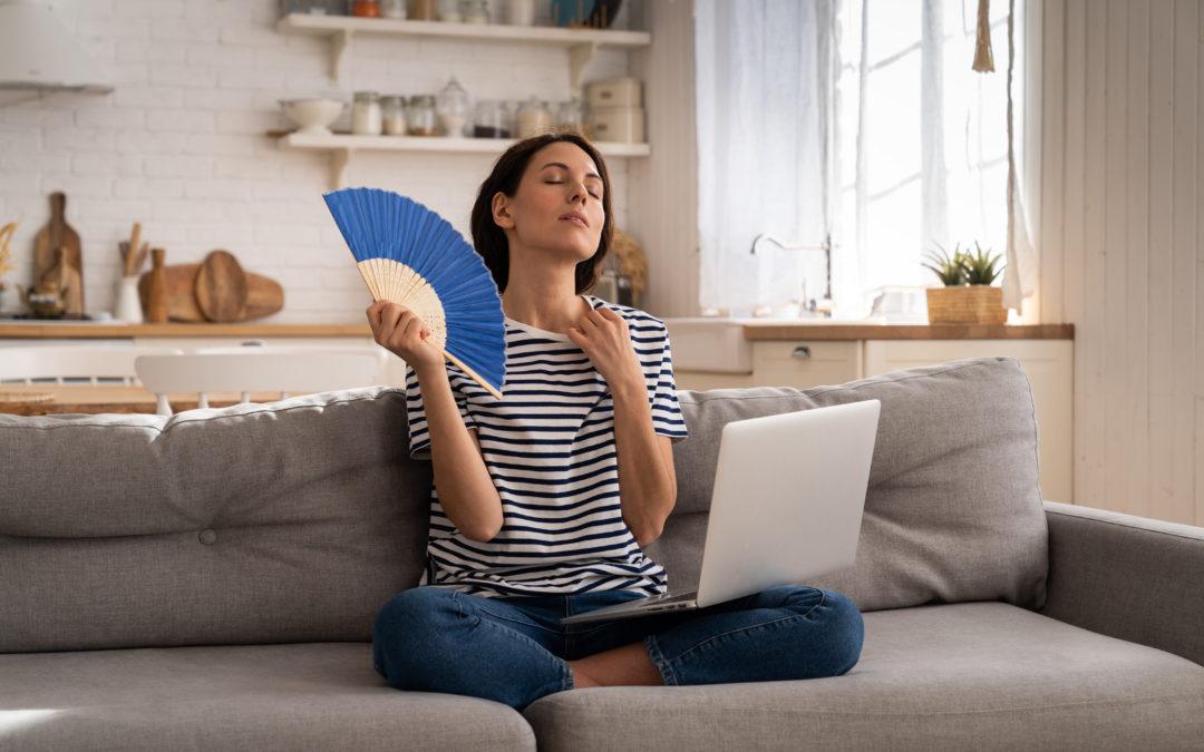 Tired millennial woman suffers from stuffiness and an inoperative air conditioner, waving blue fan sitting on couch at home working on laptop computer.