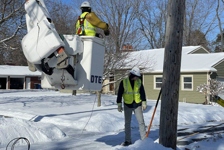 two DTE workers near a utility pole in front of house. Snow on the ground. One man in a DTE bucket lifted a few feet off the ground. The other man staring down at the pole.