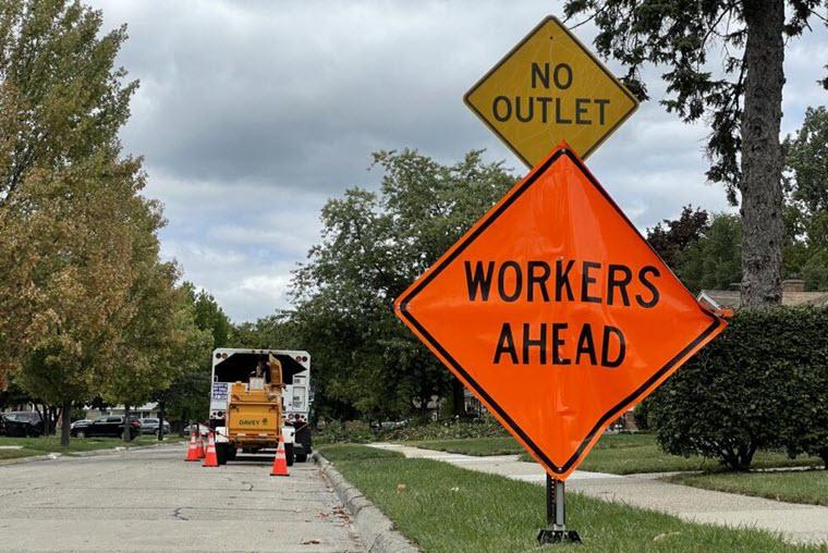 neon orange WORKERS AHEAD sign under yellow NO OUTLET sign in a neighborhood behind a tree trim truck