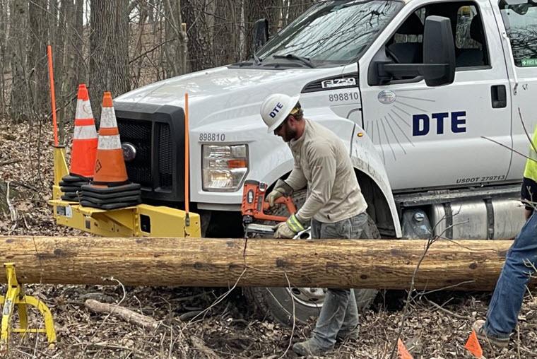 man wearing DTE hard hat, in front of DTE truck in the woods, using a power tool on a utility pole lying down