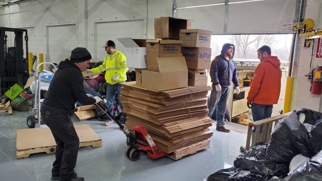 three men bringing in a stack of boxes into a warehouse